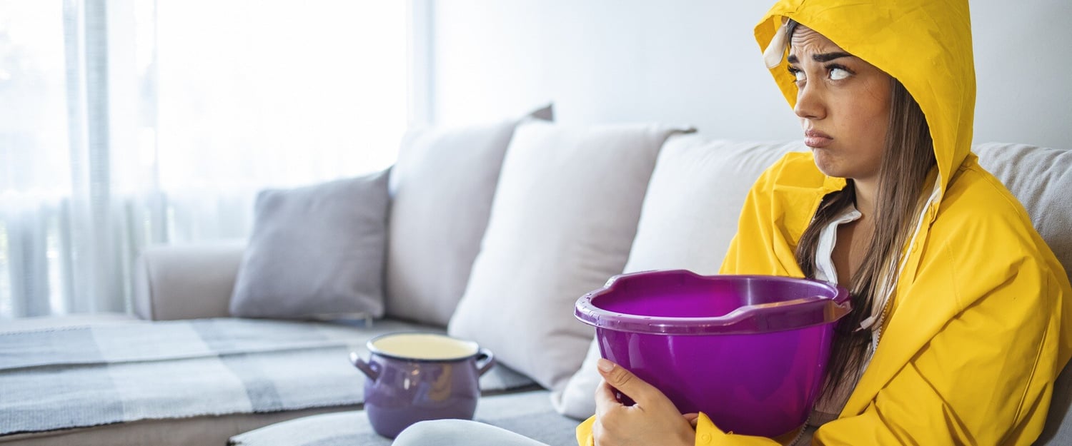 Frau sitzt mit dem Regenmantel auf ihrem Sofa und hält einen Eimer in der Hand.